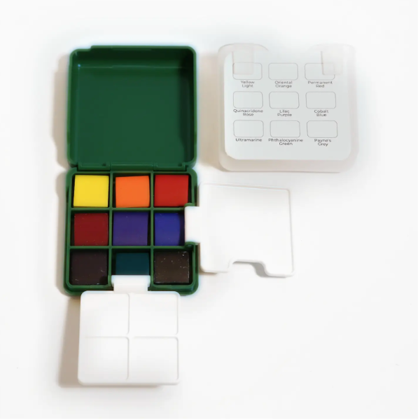 A small portable color palette set on a white background, showing a green case with multiple paint or ink wells, a white labeled reference card, and white plastic covers.