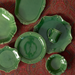 A coordinated green ceramic tableware set arranged on a red patterned fabric background, including plates, a bowl, and a mug with small metallic rivet details.
