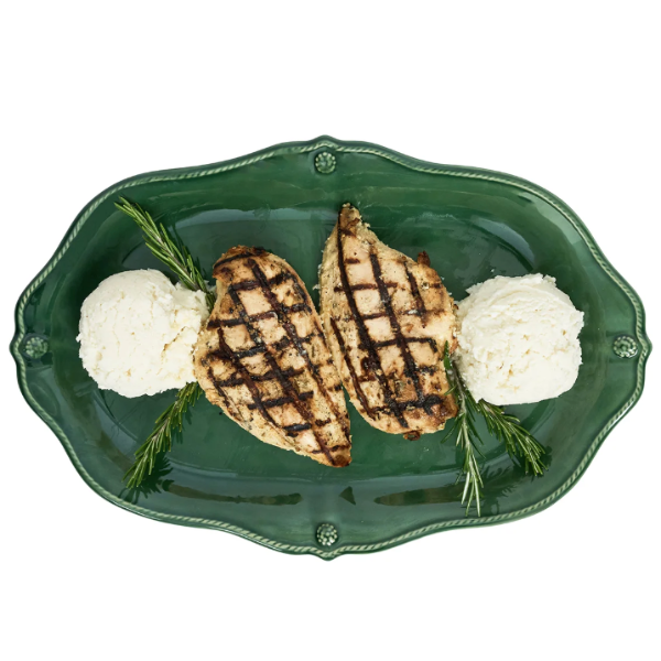 A top-down view of a green scalloped ceramic platter holding two grilled chicken breasts with white mashed potatoes and rosemary sprigs, isolated on a white background.