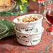 Two stacked white ceramic bowls with green winter landscape illustrations, filled with pistachios, set on a patterned red and blue table runner with wine glasses and plates in the background.