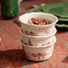 Three stacked white ceramic bowls with a red winter landscape print, filled with roasted nuts, set on a patterned red tablecloth.