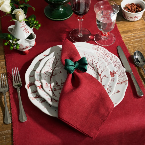 A styled holiday table setting featuring a red table runner, white plates with a red winter tree pattern, a red napkin tied with a green bow, silver cutlery, and glassware on a wooden table.