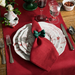 A styled holiday table setting featuring a red table runner, white plates with a red winter tree pattern, a red napkin tied with a green bow, silver cutlery, and glassware on a wooden table.