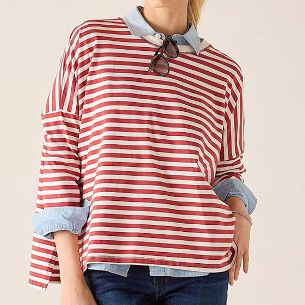 A woman wearing a red and white striped long-sleeve top with a light blue collared shirt underneath, paired with dark blue jeans, against a plain beige background.