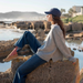 A woman sits on a large rock by the ocean wearing a light gray knit sweater, blue jeans, pink socks, tan sneakers, and a navy baseball cap, with rocky shoreline and water in the background.
