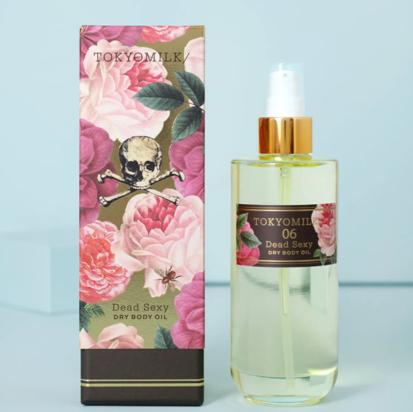 A bottle of TOKYOMILK Dead Sexy Dry Body Oil next to its illustrated packaging box, featuring pink roses, green leaves, and a skull graphic on a light blue background.