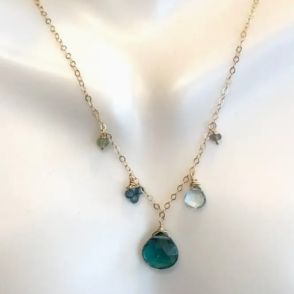 A delicate gold chain necklace displayed on a white mannequin bust. The necklace features a large, faceted green teardrop pendant at the bottom, with smaller floral and teardrop charms in green, blue, and clear stones along the chain.