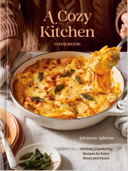 Front cover of the cookbook 'A Cozy Kitchen' by Adrianna Adarme, featuring a large photo of cheesy baked pasta in a white casserole dish with a hand lifting a spoonful. The title is in large cream text at the top, and the author name is at the bottom.
