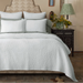 A neatly made bed in a styled bedroom setting, featuring a textured white quilt and matching white pillows with sage green side borders. The background includes a patterned headboard, a dark carved nightstand with a green patterned lamp, and a tiled floor.