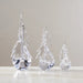 Three clear crystal Christmas tree ornaments in different sizes, arranged on a light marble surface against a neutral beige wall.