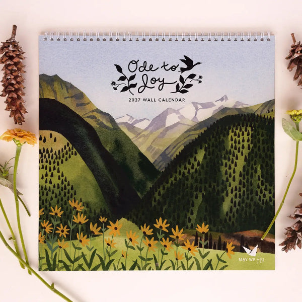 A 2027 wall calendar titled 'Ode to Joy' featuring a watercolor mountain landscape with yellow wildflowers, styled in a flat-lay with pinecones and flowers on a light beige background.