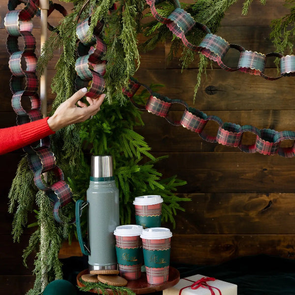 A festive holiday scene with a hand adjusting a plaid paper garland against a dark wood wall, with insulated bottles and cups on a wooden tray in the foreground.