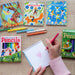 Colorful children's sketchbooks and pencils on a wooden surface with a hand drawing.