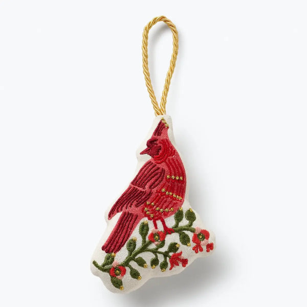 An embroidered Christmas ornament shaped like the state of Maine, featuring a red cardinal on a branch with green leaves and red berries, hanging from a gold-colored twisted string on a white background.