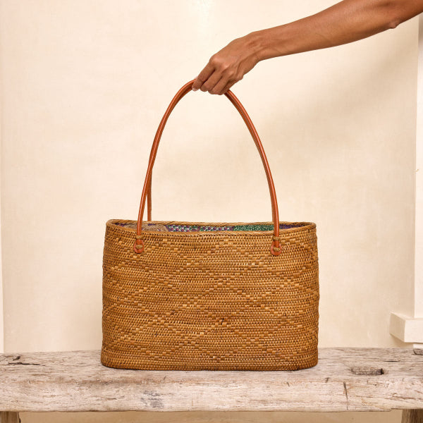Woven basket with leather handle held by a hand on a wooden surface.