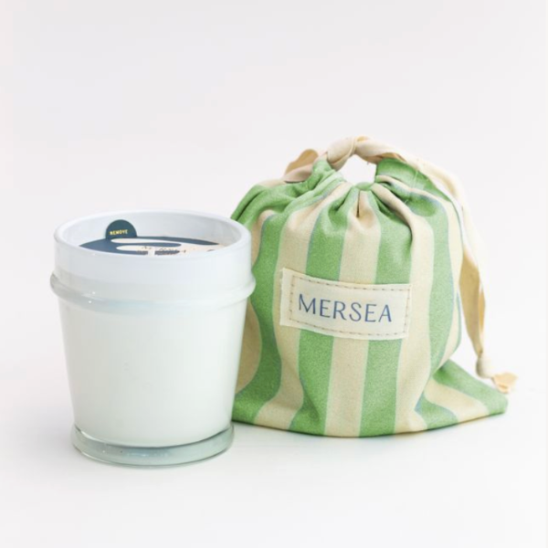 White candle in a glass jar with a green and white striped drawstring bag labeled 'MERSEA' on a light gray background.