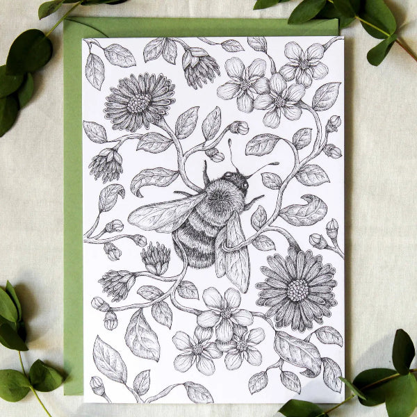 A square art print or card featuring a black-and-white detailed illustration of a bee among flowers and leaves, placed on top of a light green envelope or backing board and surrounded by green leaves on a light fabric background.