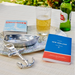 Table setting with a bottle of beer, glass, book, and anchor-shaped bottle opener on a map.