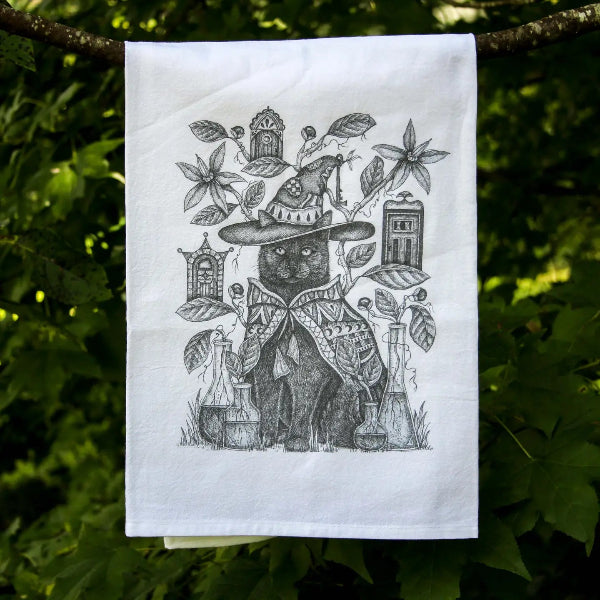 A white tea towel with a detailed black ink illustration of a witch cat wearing a hat and coat, surrounded by leaves, potion bottles, and small houses, hanging from a tree branch in a forest.