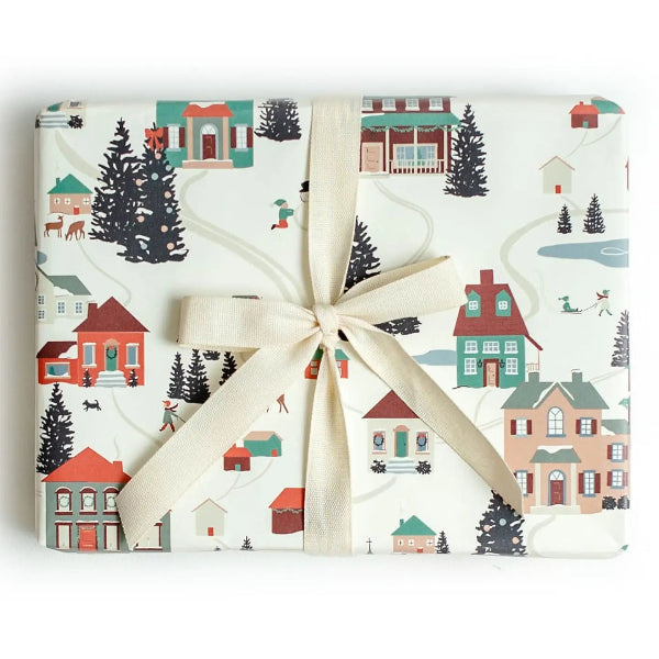 A gift-wrapped rectangular package tied with a cream ribbon bow, featuring a winter village print with houses, trees, and small figures on an off-white background.