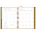 An open spiral-bound planner showing the January 2027 calendar spread. The pages have a white background with gold and brown accents, including a green and gold left margin, a gold center binding strip, and a brown right margin with monthly tabs.