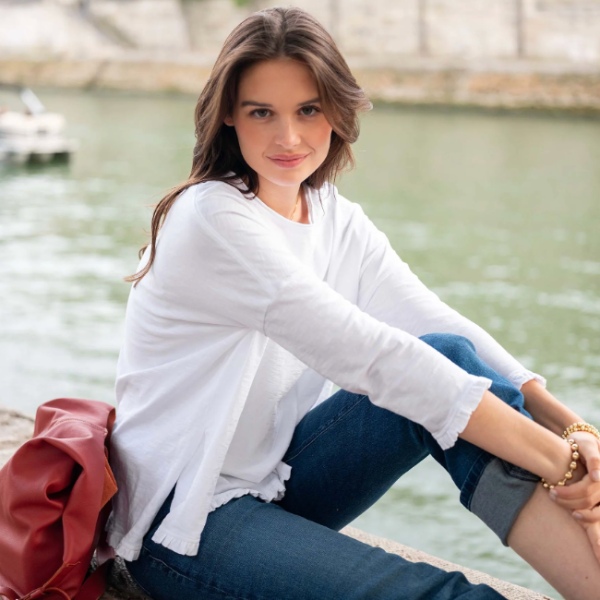 A woman sits on a stone ledge by the water, wearing a white long-sleeve top and blue jeans. A red bag rests beside her, with a blurred waterfront background.