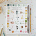 A spiral-bound watercolor painting guide book titled 'learn to paint with watercolor' lies on a white surface surrounded by colorful confetti, a paintbrush, and colored pencils. The cover features watercolor illustrations of party themes like cakes, balloons, and a swan, alongside a grid of watercolor swatches.