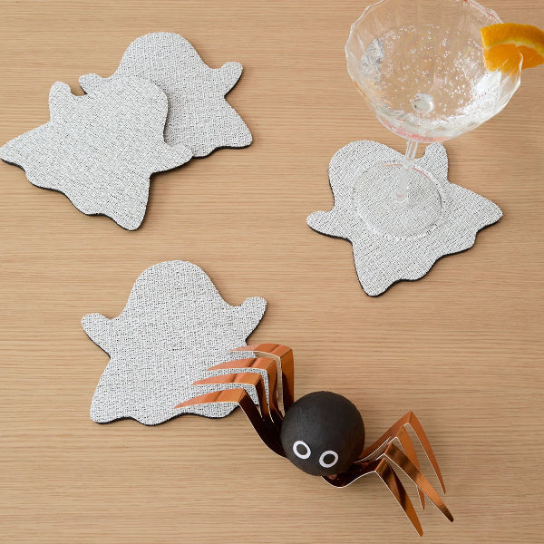 Halloween-themed coasters shaped like ghosts and a spider, arranged on a light wood tabletop next to a cocktail glass with a garnish.
