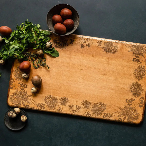 Wooden cutting board with floral patterns surrounded by fresh herbs and eggs on a dark surface