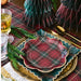 A styled holiday table setting featuring a scalloped ceramic plate with a red and green tartan plaid pattern and gold trim, layered on top of a matching green plaid plate with a gold charger plate underneath. The setting includes gold flatware, a glass of amber beverage, a glass of water, and pine greenery, with decorative paper trees in the background.