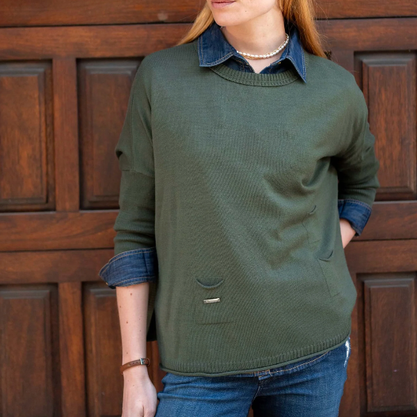 A woman wearing an olive green knit sweater layered over a blue denim collared shirt and blue jeans, standing in front of a dark brown wooden paneled door.