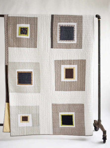 A geometric patchwork quilt draped over a black metal stand, featuring blocks of taupe, cream, navy blue, and mustard yellow.