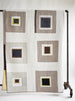 A geometric patchwork quilt draped over a black metal stand, featuring blocks of taupe, cream, navy blue, and mustard yellow.