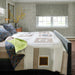 A styled bedroom interior showing a bed made with a patchwork quilt in cream, beige, and brown tones, blue floral pillows, and a bright green folded throw. The room has patterned wallpaper, a window with gray roman blinds, and a dark wood floor.