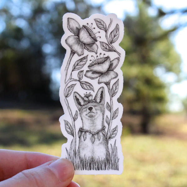A hand holds a die-cut sticker featuring a black and white sketch of a dog surrounded by flowers, with a blurred outdoor park background.