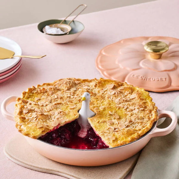 Pie in a pink Le Creuset dish with a seagull pie weight on a light pink surface.