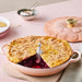 Pie in a pink Le Creuset dish with a seagull pie weight on a light pink surface.