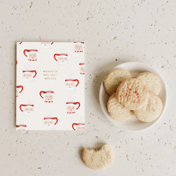 A flat-lay of a white greeting card with a Santa Claus pattern and a plate of sugar cookies on a textured light gray surface.