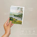 Hand holding a plantable seed paper envelope with a scenic design on a beige background.