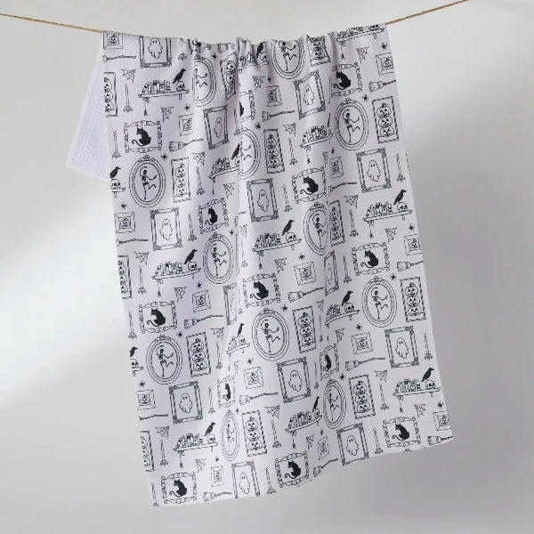 A white kitchen tea towel with a black whimsical pattern of cats, birds, and framed illustrations, hanging from a string on a light gray background.
