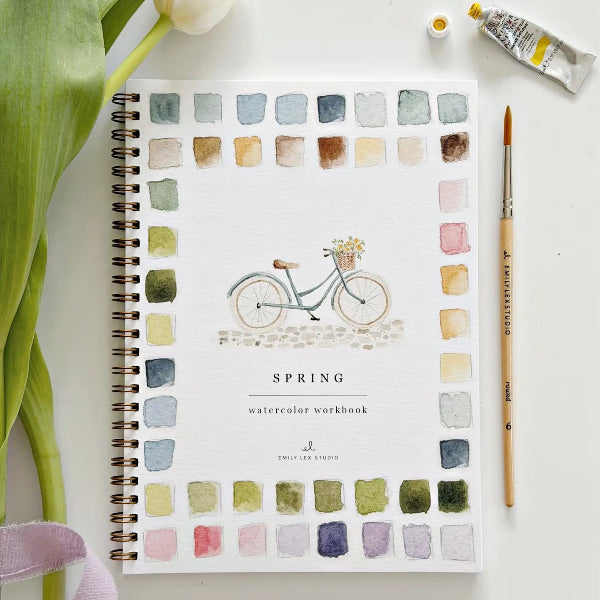 Spring watercolor workbook with a bicycle illustration and color swatches, surrounded by art supplies on a white surface.