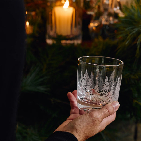 A hand holds a clear glass rocks tumbler decorated with an etched winter forest scene of pine trees, set against a dark, festive background with pine branches and a glowing candle.