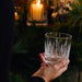 A hand holds a clear glass rocks tumbler decorated with an etched winter forest scene of pine trees, set against a dark, festive background with pine branches and a glowing candle.