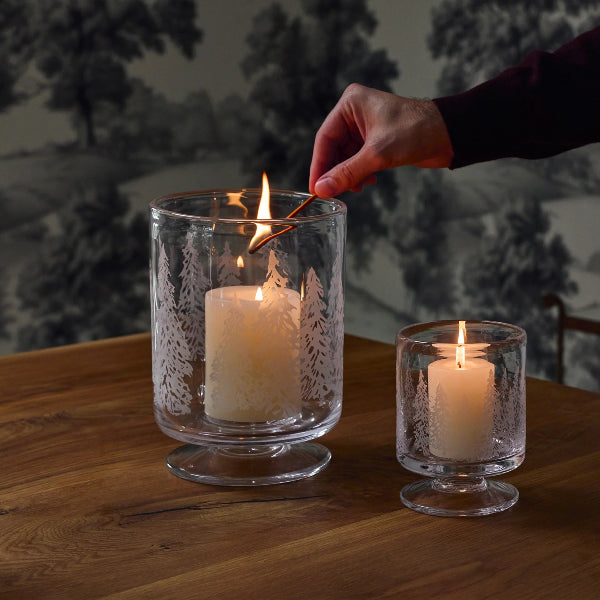 Two clear glass candle holders with etched tree patterns sit on a wooden table, one lit with a white pillar candle and the other with a smaller candle. A hand holds a match above the larger candle.