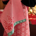 A close-up of a red and white gingham kitchen towel with a green border featuring a red house and white snowflake pattern, held in front of a pink floral apron.