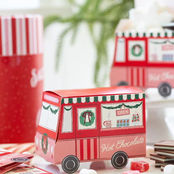 A small red and pink hot chocolate truck-shaped box sits on a tabletop with candy pieces nearby. A larger red and white striped hot chocolate container is blurred in the background, along with a second hot chocolate truck box and a soft holiday-themed setting.