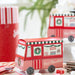 A small red and pink hot chocolate truck-shaped box sits on a tabletop with candy pieces nearby. A larger red and white striped hot chocolate container is blurred in the background, along with a second hot chocolate truck box and a soft holiday-themed setting.