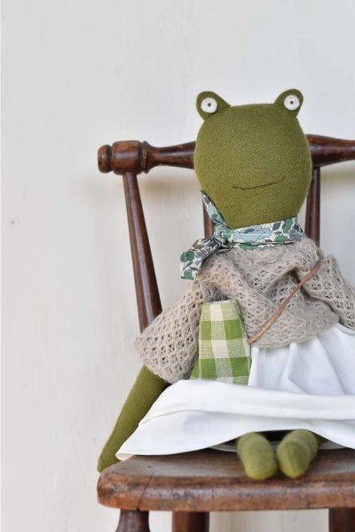 Frog-shaped plush toy wearing a dress with a green plaid pattern, sitting on a wooden chair.