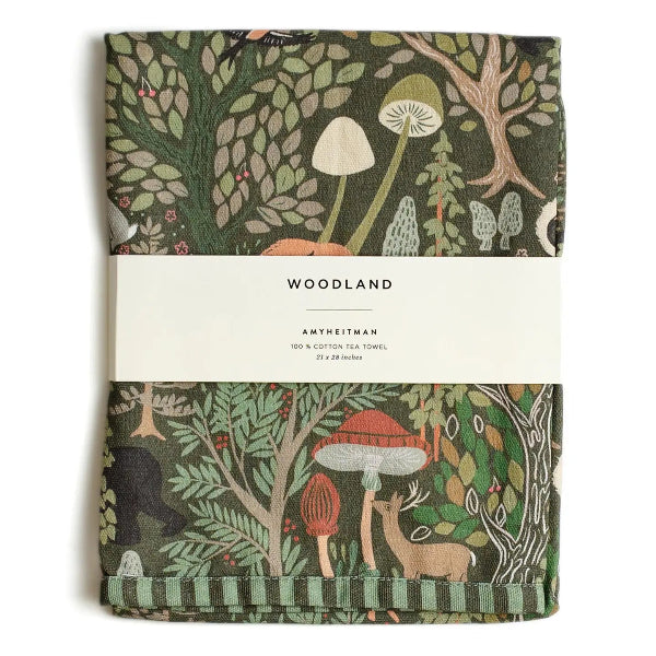 A folded cotton tea towel with a woodland forest pattern, wrapped in a white paper band labeled 'WOODLAND AMYHEITMAN'. The pattern features green trees, mushrooms, and a deer.