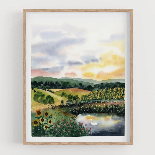 A framed watercolor landscape art print featuring a serene countryside scene with rolling green hills, a pond with a white swan, sunflowers, and a cloudy sunset sky, presented in a light wood frame against a white wall.